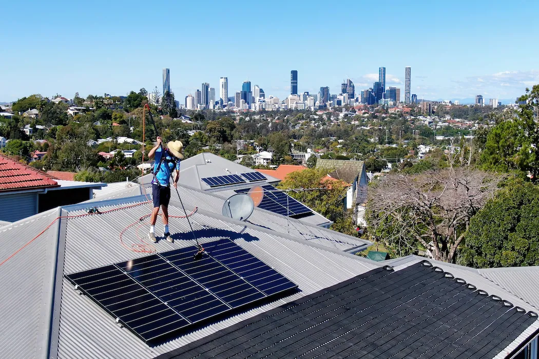 kg-roof-cleaning-2 Kleen Genie technician standing on roof cleaning solar panels with equipment and Brisbane city on the horizon